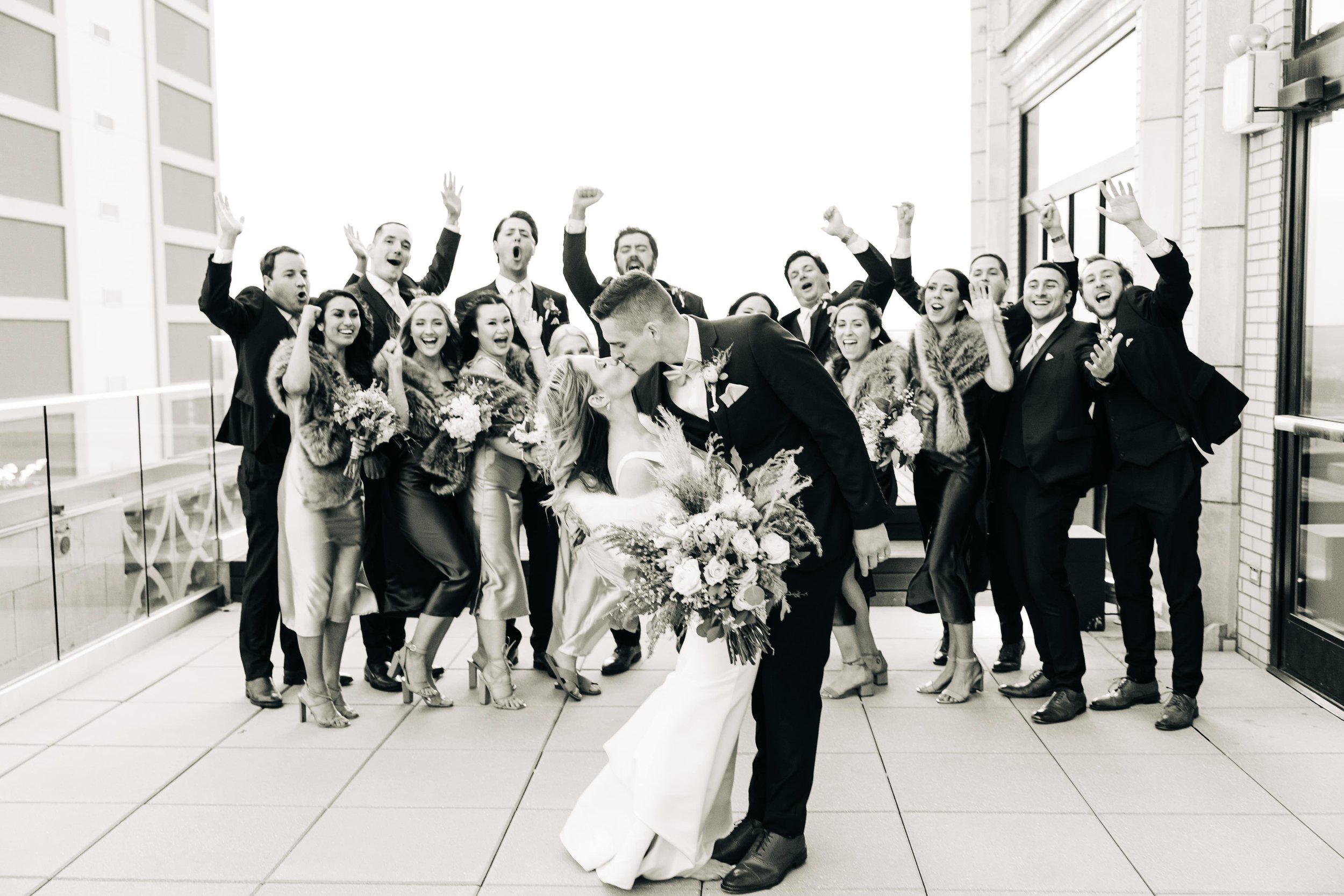 Black and white photo of a wedding party cheering while the bride and groom kiss on the Starlight Terrace at Hotel Kansas City